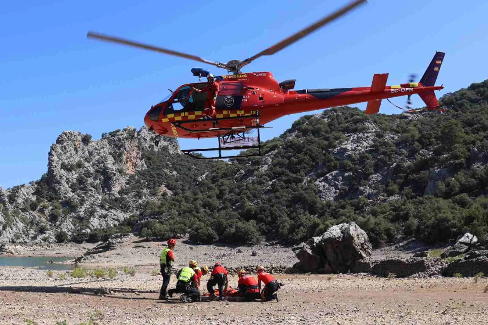 Un helicóptero rojo de los Bomberos de Mallorca sobrevolando un área montañosa mientras varios efectivos atienden a una persona en el suelo