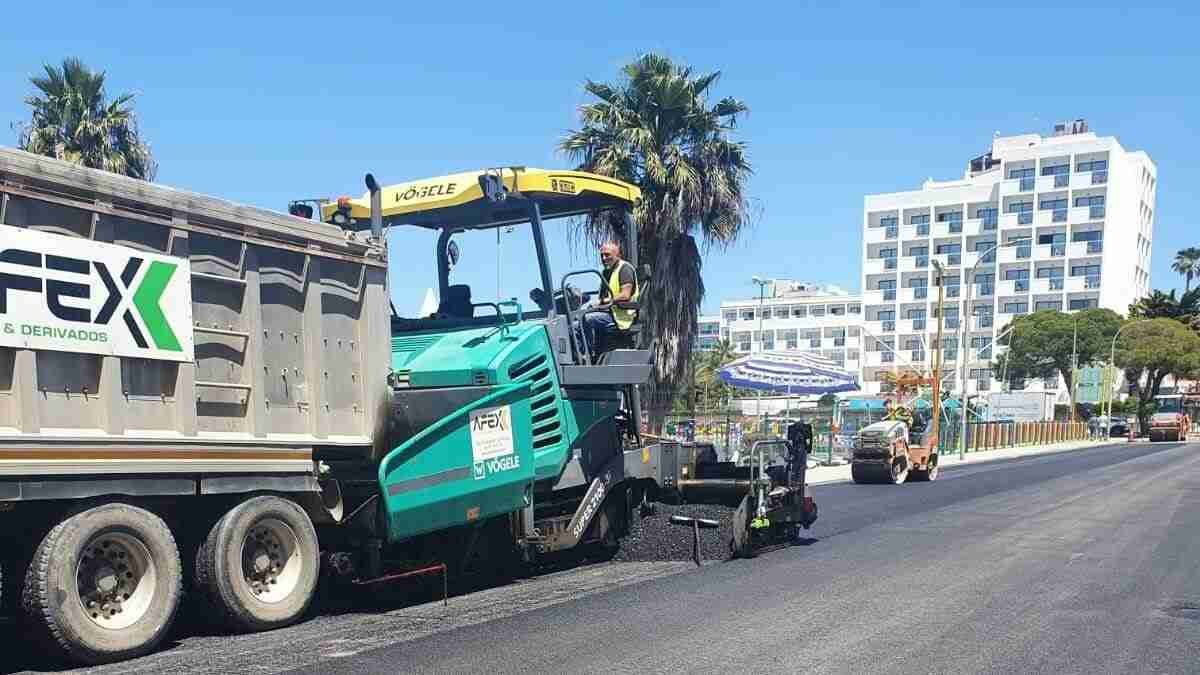 Una gran máquina extendedora de asfalto de color verde y un camión de la empresa Fex operan sobre el nuevo pavimento negro de una avenida en Cala Millor bajo un cielo despejado