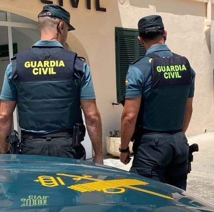 Dos agentes de la Guardia Civil de espaldas portando chalecos antibalas con las letras amarillas del cuerpo médico y policial junto al capó de un coche oficial en Mallorca