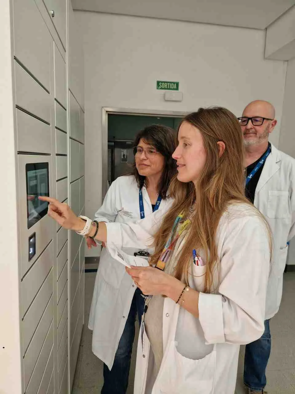Tres profesionales sanitarios con bata blanca comprobando el funcionamiento de una pantalla táctil integrada en un gran mueble de taquillas automatizadas de color blanco