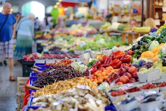 Gran variedad de frutas, cerezas, pimientos y verduras expuestas en cajas en un mercado público con clientes al fondo