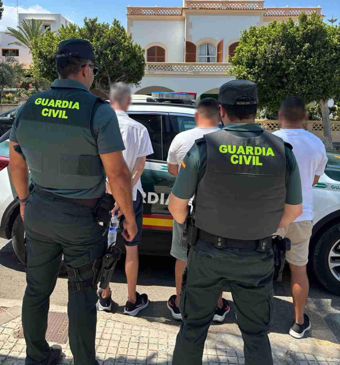 Dos agentes de la Guardia Civil custodiando a tres detenidos de espaldas junto a un coche patrulla en Felanitx