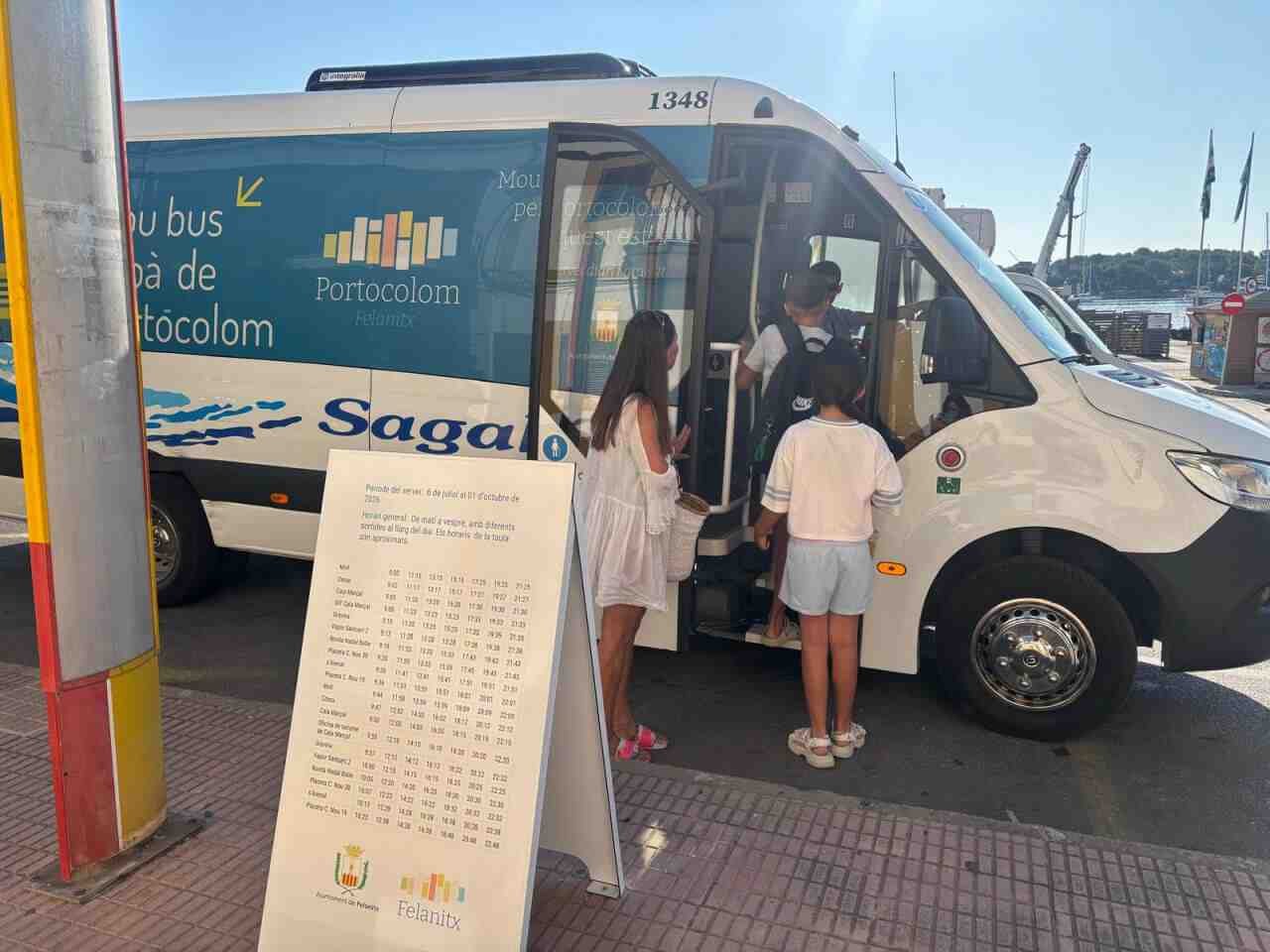Usuarios y niños subiendo al nuevo minibús urbano gratuito de Portocolom junto al cartel de horarios oficiales