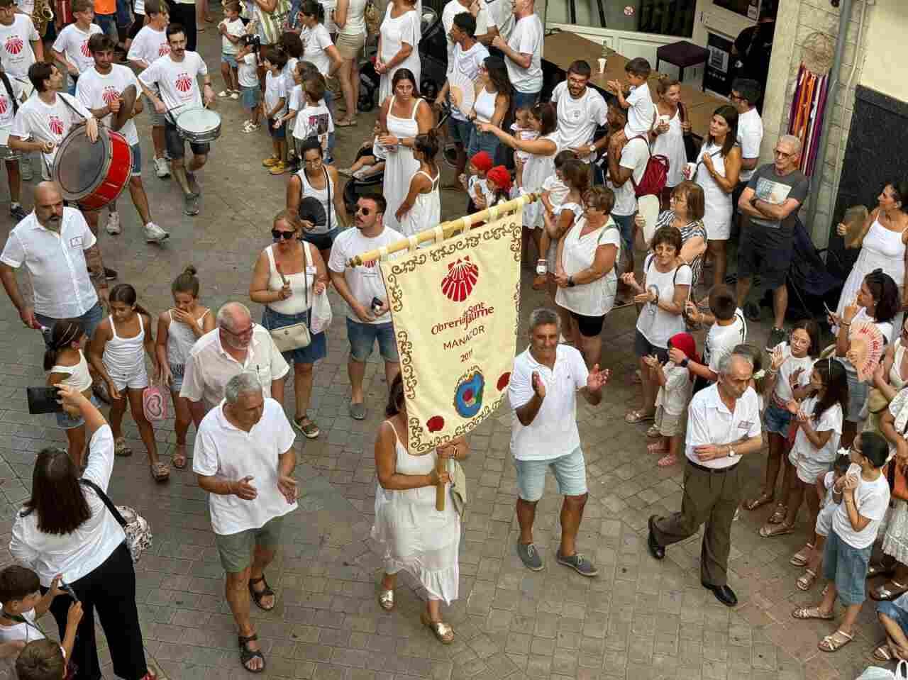 Vecinos vestidos de blanco desfilan en la Crida de Sant Jaume de Manacor portando un estandarte de la Obreria
