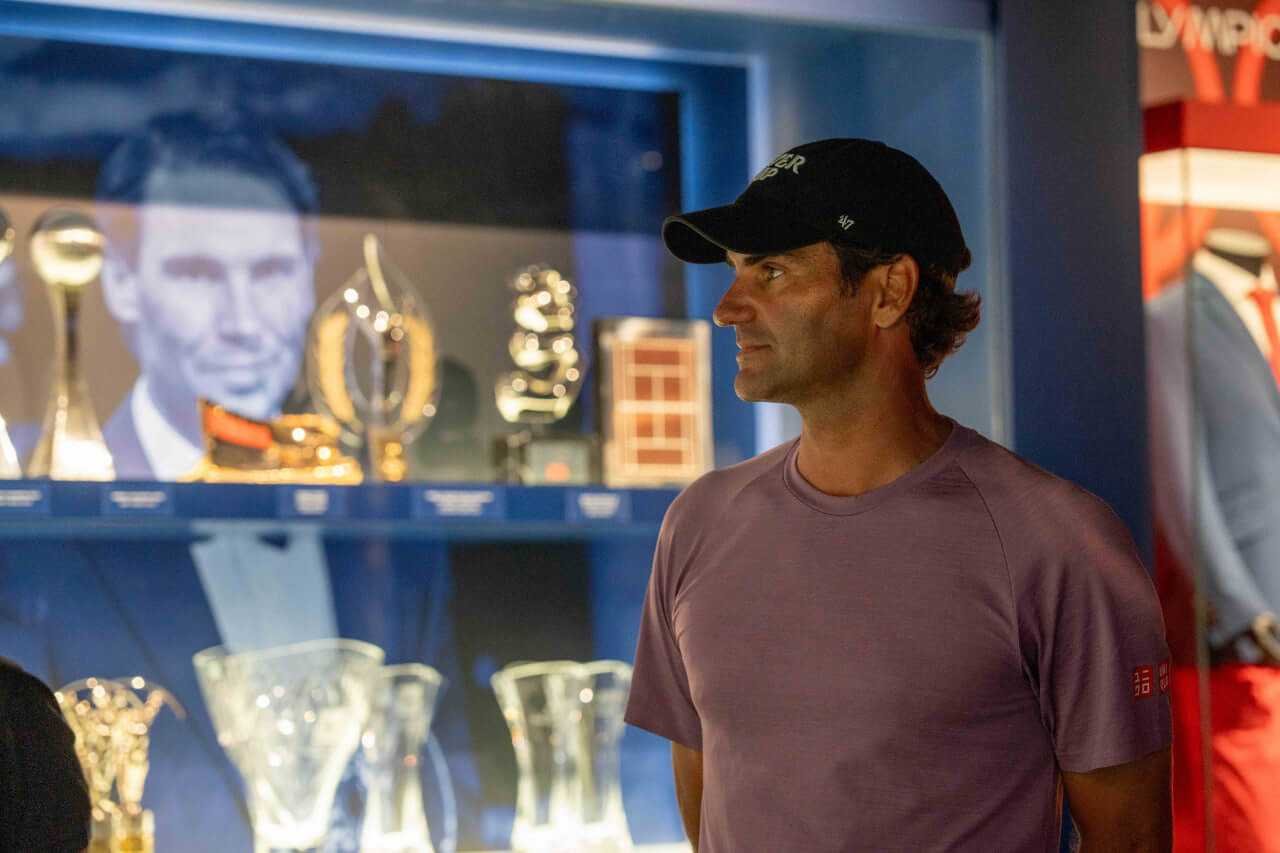 Roger Federer con gorra oscura y camiseta violeta observando los trofeos del Rafa Nadal Museum