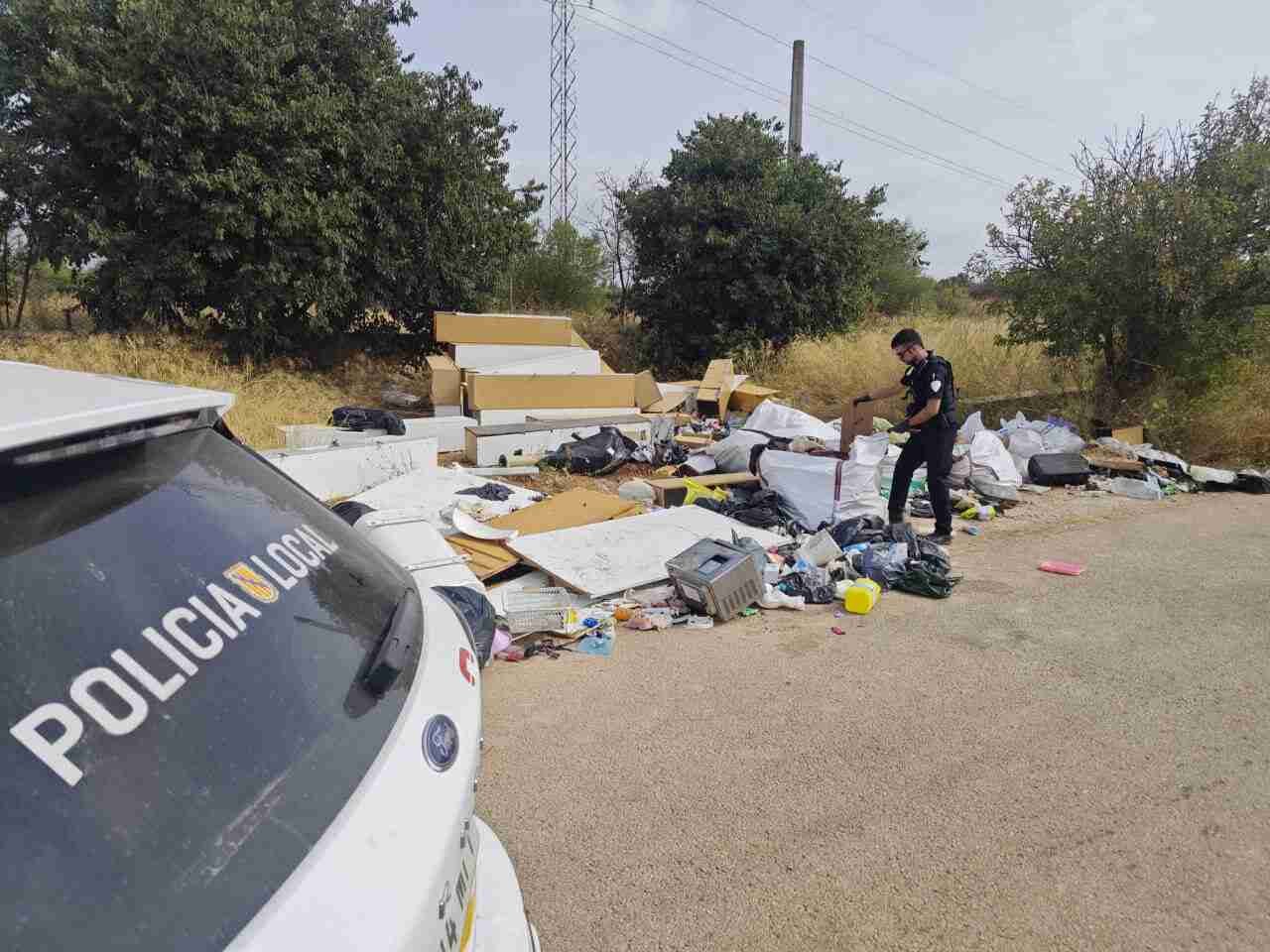 Agente de policía local revisando enseres y basura abandonados junto a un vehículo patrulla