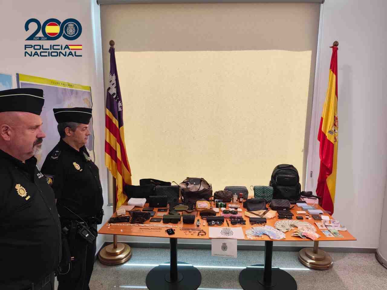 Dos agentes de la Policía Nacional junto a una mesa con bolsos joyas y dinero recuperados