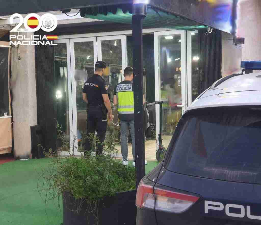 Dos agentes de la Policía Nacional de espaldas frente a un local acristalado junto a un coche patrulla durante una detención