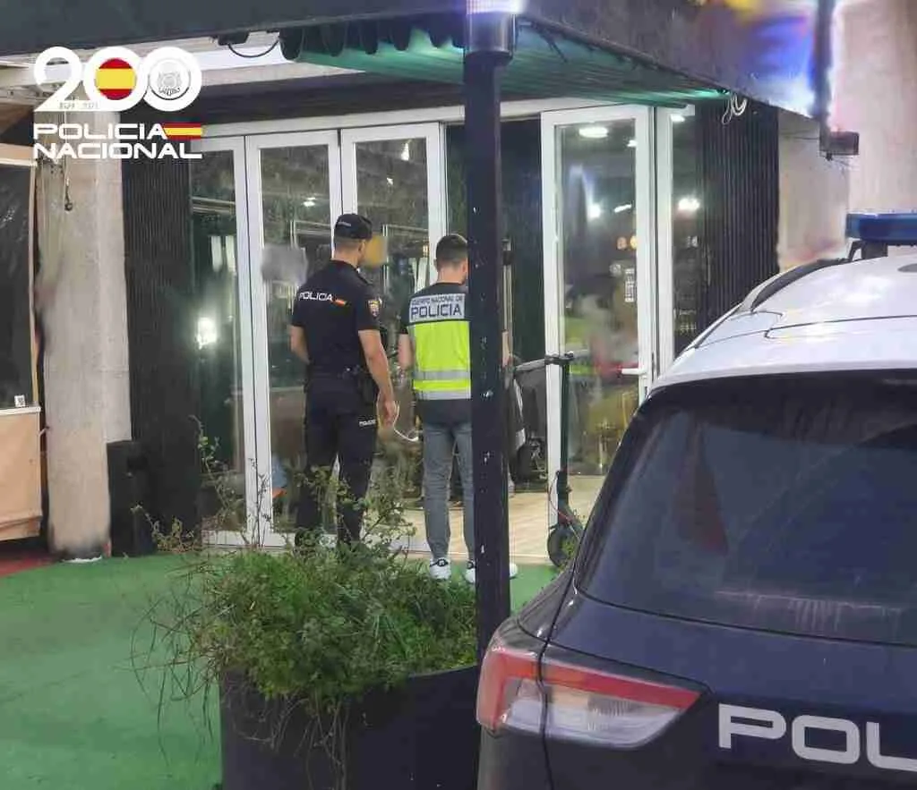 Dos agentes de la Policía Nacional de espaldas frente a un local acristalado junto a un coche patrulla durante una detención