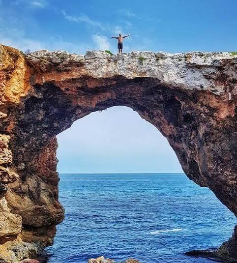 Persona con los brazos abiertos en lo alto del puente de roca natural sobre el mar en Cala Varques