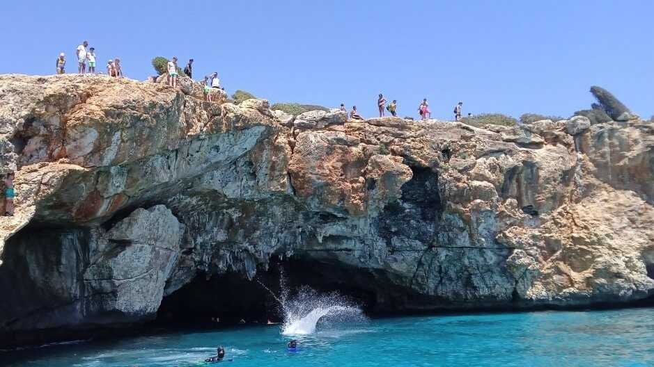 Multitud de personas sobre los acantilados de roca y salto al agua frente a la cueva en Cala Varques