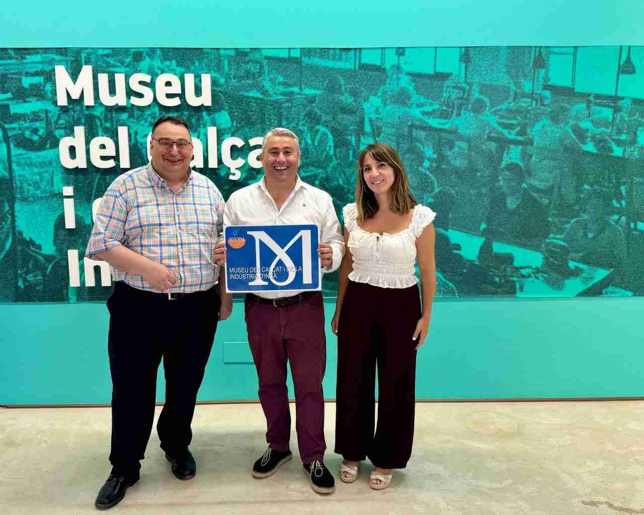 Tres personas posan sonrientes sosteniendo la placa acreditativa del ICOM en el Museu del Calçat i de la Indústria d’Inca frente a un mural verde