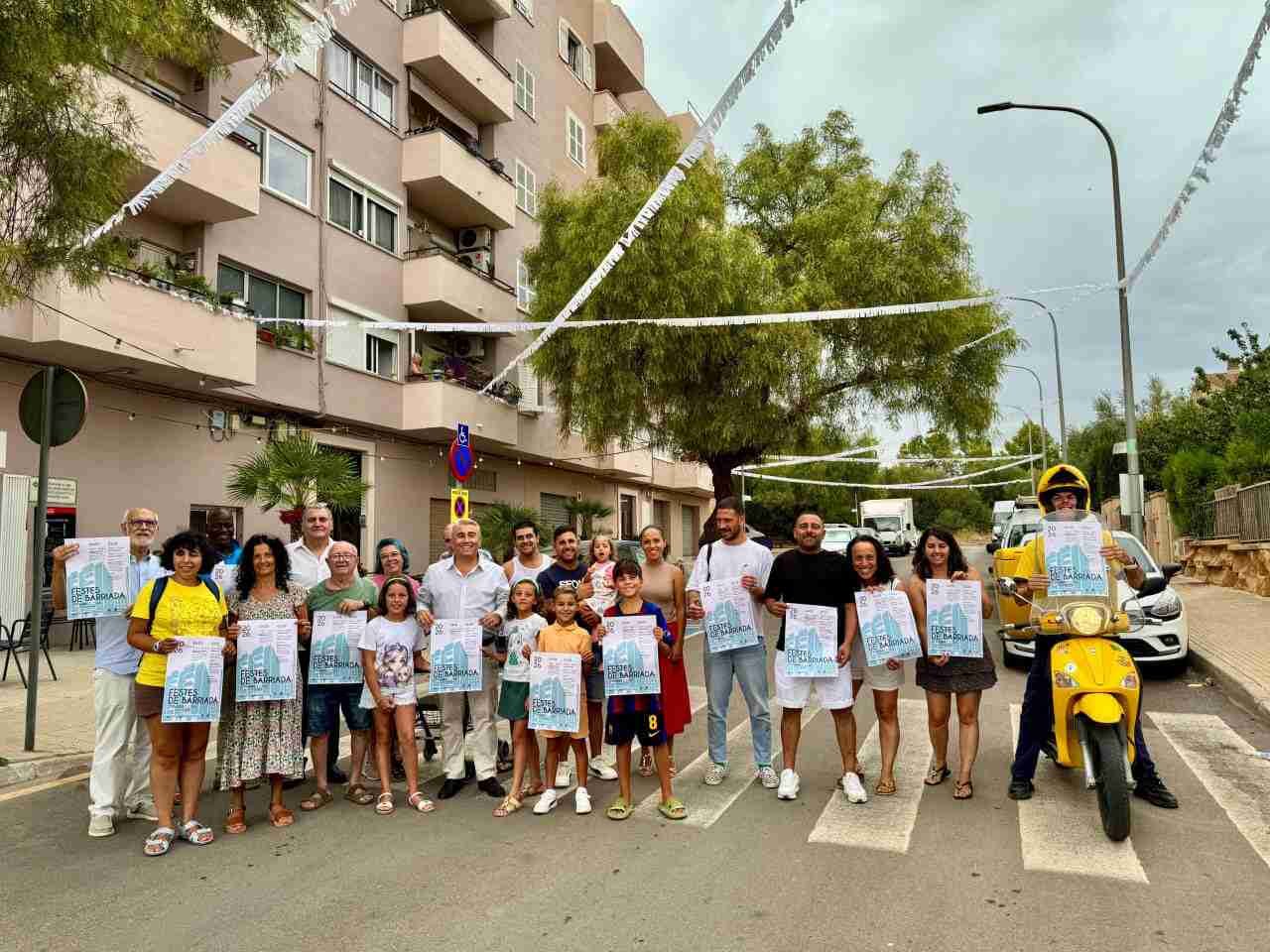 Vecinos y representantes municipales posan en la calle con el cartel festivo de Fernández Cela junto a una moto amarilla