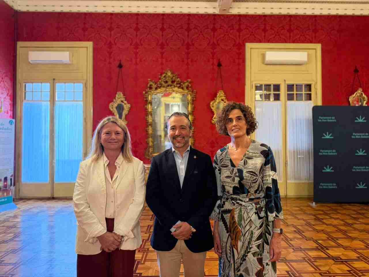 Núria Riera, Llorenç Galmés y una diputada posan de pie en un salón del Parlamento Balear con paredes rojas damascadas